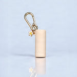 A beige cylindrical cosmetic case with a gold carabiner clip and a small white flower charm with yellow accents, standing on a reflective light blue surface.