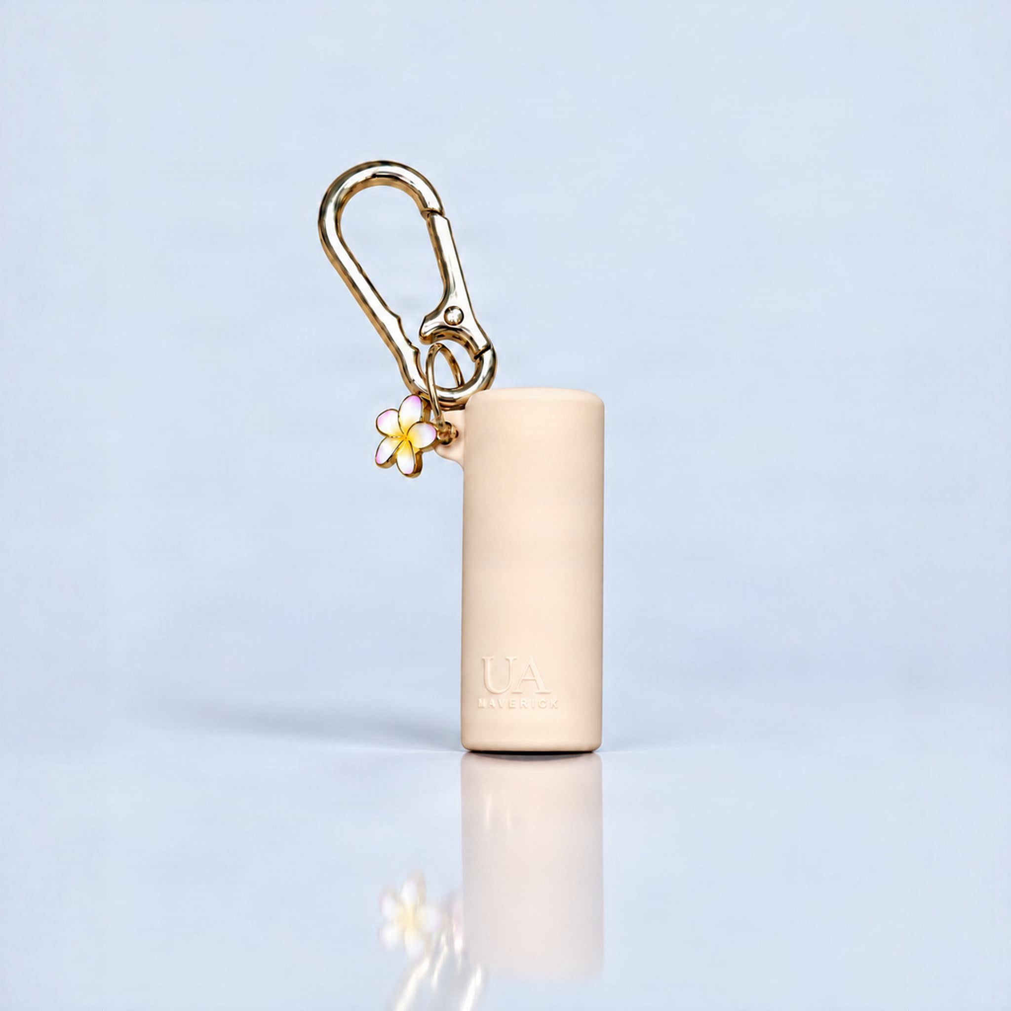 A beige cylindrical cosmetic case with a gold carabiner clip and a small white flower charm with yellow accents, standing on a reflective light blue surface.
