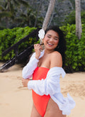 A woman on a sandy beach wearing a bright coral-red one-piece swimsuit with a white long-sleeve shirt draped off her shoulders, holding a white flower.