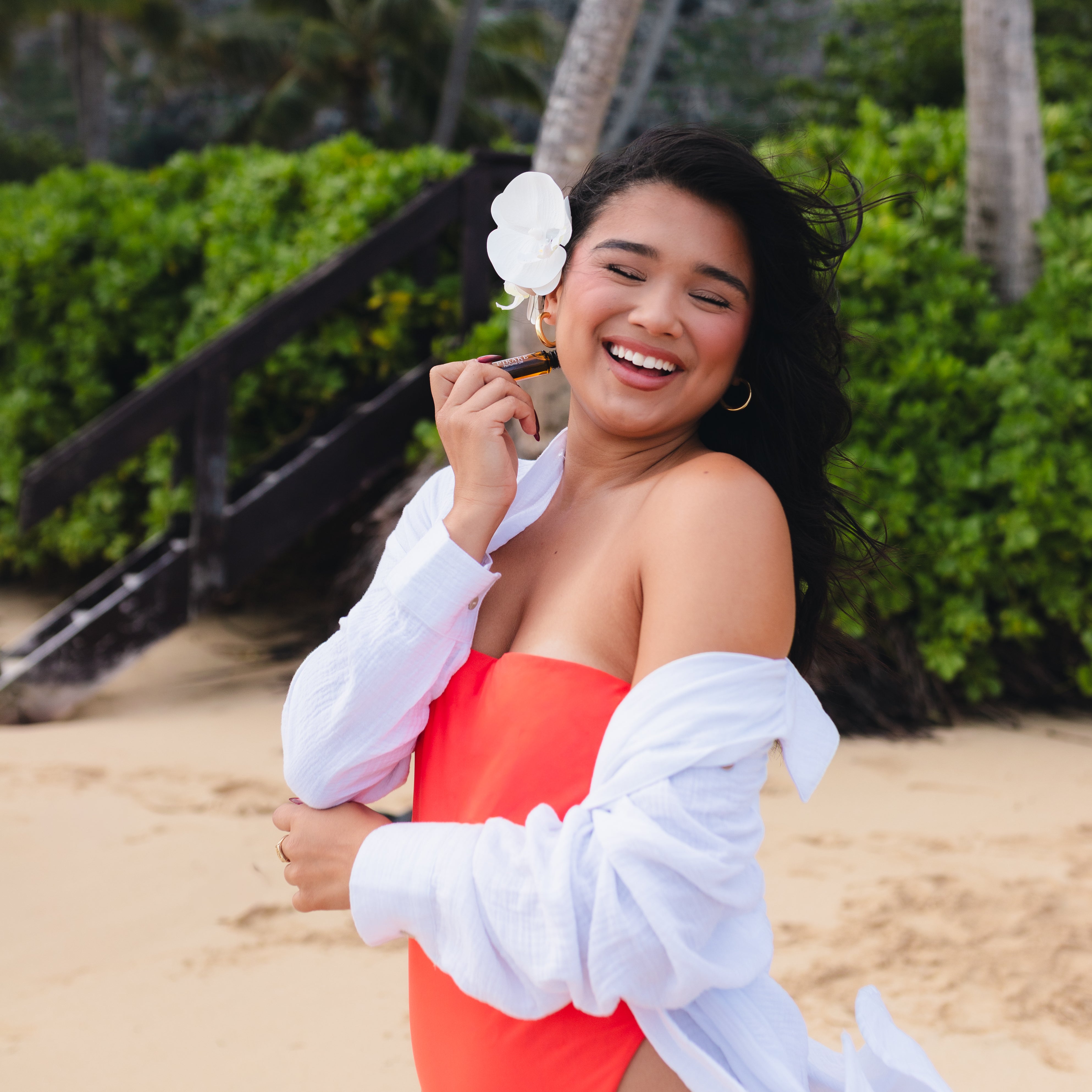 A woman on a sandy beach wearing a bright coral-red one-piece swimsuit with a white long-sleeve shirt draped off her shoulders, holding a white flower.